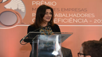 Foto da Prof.ª Lúcia França (Primeira-dama do estado de São Paulo) em pé, apoiada no púlpito de acrílico, falando ao microfone. Lúcia veste botas de cano alto, uma saia longa na cor verde, blusa estampada e um casaquinho curto na cor preta.