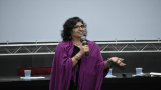 Foto da palestrante Andrea Parra falando ao microfone, em pé, no palco.