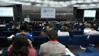 Foto do auditório da Secretaria. Em primeiro plano, pessoas sentadas em poltronas azuis. Ao fundo, a palestrante Andrea Parra falando ao microfone, em pé.