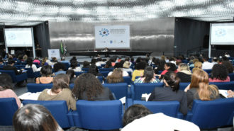 Foto ampliada do auditório da Secretaria. Em primeiro plano pessoas sentadas nas poltronas azuis do auditório e à frente, na mesa o Prof. Luiz Davi Alberto Araújo falando ao microfone.