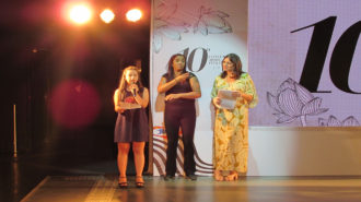 Foto de uma das modelos falando ao microfone. Ao fundo, a apresentadora do evento e a intérprete de Libras.