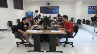 Foto de usuários sentados em cadeiras pretas, em frente aos computadores. Ao fundo, o banner do Programa POETA e uma técnica em pé.
