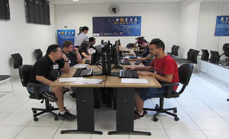 Foto de usuários sentados em cadeiras pretas, em frente aos computadores. Ao fundo, o banner do Programa POETA e uma técnica em pé.