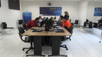 Foto de usuários sentados em cadeiras pretas, em frente aos computadores. Uma técnica está em pé, ao lado dos usuários.