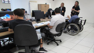 Foto da sala de aula. Em primeiro plano, usuários sentados em cadeiras pretas, em frente ao computador, olhando para apostilas. Ao fundo, em pé, duas técnicas.