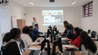 Foto da sala de aula. Em primeiro plano, usuários sentados em frente aos computadores. Ao fundo, um intérprete de Libras, em pé, e uma técnica próxima a um usuário.