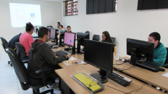 Foto de usuários sentados em frente aos computadores. Ao fundo, uma técnica sentada em frente ao computador.