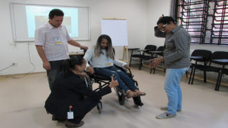 Foto de uma usuária sentada em uma cadeira de rodas preta, dois usuários em pé e uma técnica abaixada perto da cadeira.