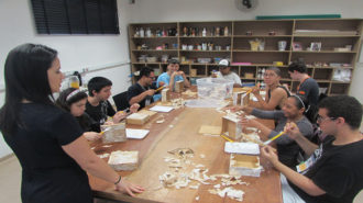 Foto da sala de artes do Centro de Tecnologia e Inovação. Nove usuários sentados próximos a uma mesa grande e uma técnica em pé. Os usuários estão colando pedaços de papel em caixas pequenas.