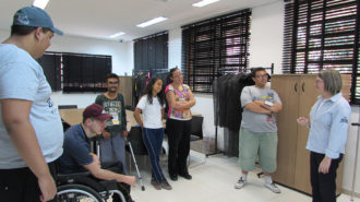 Foto da sala de aula. Cinco usuários em pé e um na cadeira de rodas conversando com a técnica.