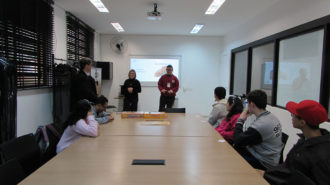 Foto da sala de aula. Seis usuários sentados próximos a uma mesa e dois técnicos e um usuário em pé. Os usuários participam de uma atividade de mímica.
