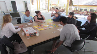 Foto do pátio do Centro de Tecnologia e Inovação. Cinco usuários, duas técnicas e um intérprete de Libras sentados em cadeiras pretas, próximas a uma mesa. Os usuários estão jogando bingo.