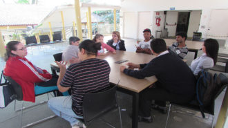 Foto do pátio do Centro de Tecnologia e Inovação. Cinco usuários, três técnicos e um intérprete de Libras sentados em cadeiras pretas, próximas a uma mesa. Os usuários estão conversando.