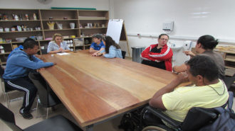 Foto da sala de artes do Centro de Tecnologia e Inovação. Seis usuários, uma técnica e um intérprete de Libras sentados em cadeiras pretas, próximas a uma mesa grande de madeira. Os usuários estão conversando.