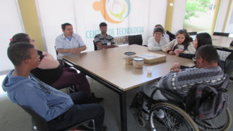 Foto do pátio do Centro de Tecnologia e Inovação. Seis usuários, uma técnica e um intérprete de Libras sentados em cadeiras pretas, próximas a uma mesa. Os usuários estão conversando.