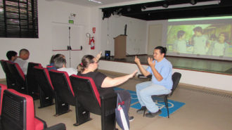 Foto do auditório do Centro de Tecnologia e Inovação. Em primeiro plano, cinco usuários sentados em poltronas vermelhas e um intérprete de Libras. Ao fundo, a exibição de um filme.