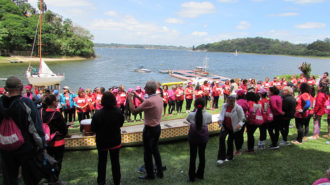 Foto da cerimônia das rosas. Remadoras Rosas reunidas ao redor de um barco Dragon Boat, segurando rosas.