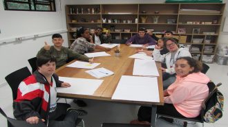 Foto da sala de artes. Nove usuários sentados próximos à mesa, desenhando em folhas brancas.