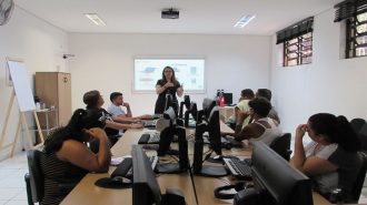 Foto da sala de aula. Em primeiro plano, 7 usuários sentados em frente aos computadores. Ao fundo, uma técnica e a lousa interativa.