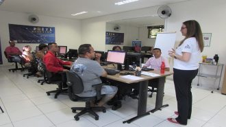 Foto da sala de aula. 7 usuários sentados em frente aos computadores e uma técnica em pé.