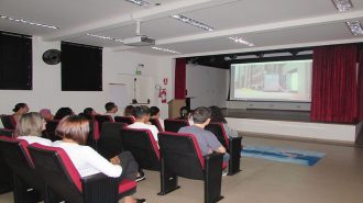 Foto do anfiteatro. Em primeiro plano, 11 usuários e duas técnicas sentados em cadeiras pretas e vermelhas. Ao fundo, o telão do auditório.