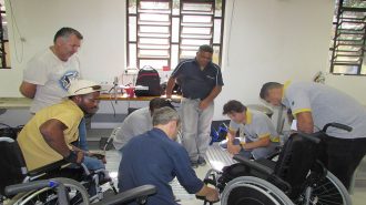Foto da oficina. Um professor mexendo em uma cadeira de rodas e sete usuários observando.