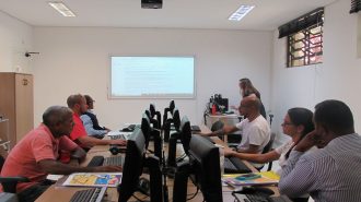 Foto do laboratório. Em primeiro plano, seis usuários sentados em frente aos computadores. Ao fundo, uma técnica olhando para o computador.