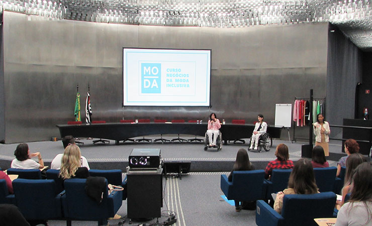 Em primeiro plano, usuários sentados em poltronas. Ao fundo, a Secretária de Estado, Célia Leão, e a Gestora do Programa Moda Inclusiva, Izabelle Palma, no palco do auditório da Secretaria de Estado dos Direitos da Pessoa com Deficiência.