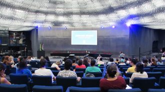 Imagem do auditório da Secretaria de Estado dos Direitos da Pessoa com Deficiência de São Paulo. Em primeiro plano, alunos sentados em poltronas. Ao fundo, no palco do auditório, a gerente do Programa Moda Inclusiva, Izabelle Palma.
