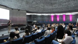 Imagem dos alunos do curso de Moda Inclusiva – Módulo Criação sentados em poltronas e da professora, em pé, falando ao microfone, no palco do auditório da Secretaria de Estado dos Direitos da Pessoa com Deficiência de São Paulo.