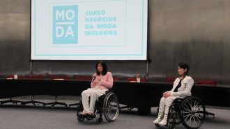 Imagem da Secretária Célia Leão, falando ao microfone, ao lado da gerente do Programa Moda Inclusiva Izabelle Palma, no palco do auditório da Secretaria de Estado dos Direitos da Pessoa com Deficiência de São Paulo.