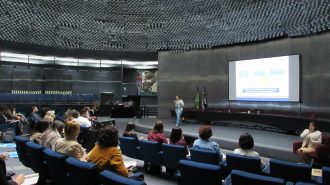 Imagem do auditório da Secretaria de Estado dos Direitos da Pessoa com Deficiência de São Paulo. Em primeiro plano, alunos sentados em poltronas. Ao fundo, no palco do auditório, a professora do curso.