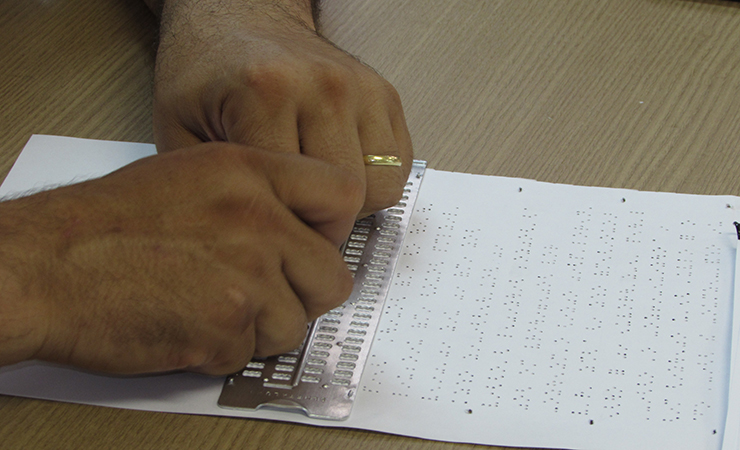 Duas mãos utilizando a reglete para escrever em Braille