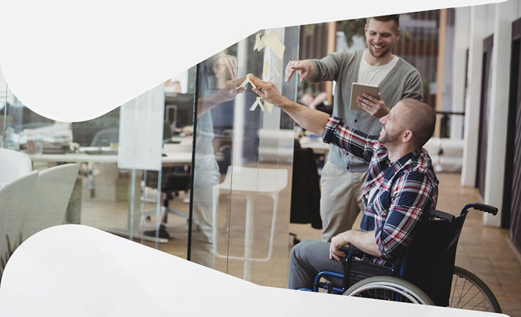 Foto de dois homens em ambiente corporativo, um deles utiliza cadeira de rodas.