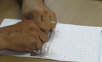 Fotografia colorida. Duas mãos escrevendo em braille em uma folha branca sobre uma mesa de madeira.