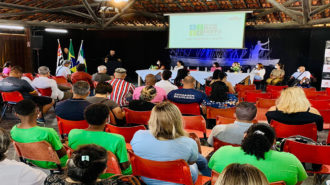 Imagem colorida do Lançamento do Programa Todas in-Rede em Ilha Comprida. Em primeiro plano pessoas sentadas em cadeiras no teatro. Ao fundo, no palco, autoridades durante pronunciamentos.