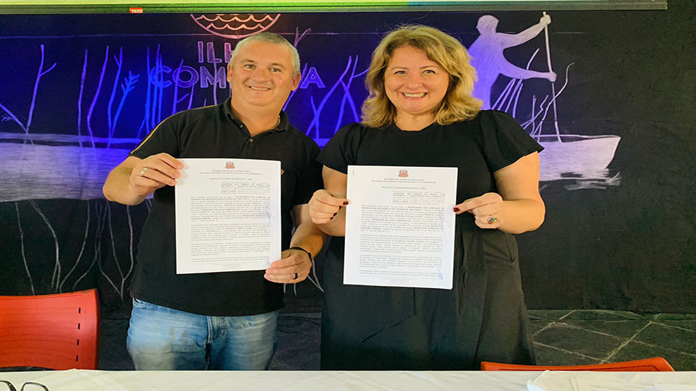 Imagem colorida do Lançamento do Programa Todas in-Rede em Ilha Comprida. A Secretária Aracélia Costa e o prefeito da cidade segurando termo de parceria.