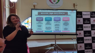 Imagem colorida do lançamento do Programa Todas in-Rede em Valinhos. A Secretária de Estado dos Direitos da Pessoa com Deficiência, Aracélia Costa, em pé, ao microfone. Ao fundo, slide do programa.