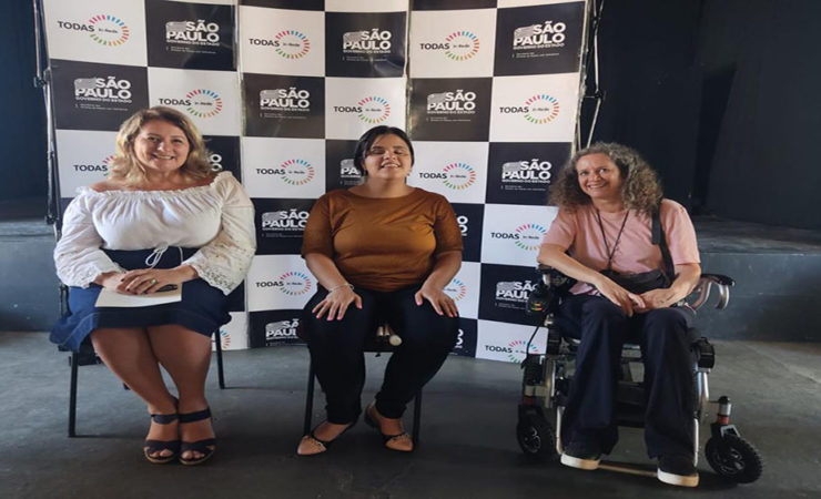 Imagem colorida do Encontro de Mulheres em Capivari – Todas in-Rede. A Secretária Arácelia Costa e outras mulheres, sentadas, em frente ao painel do programa no evento.