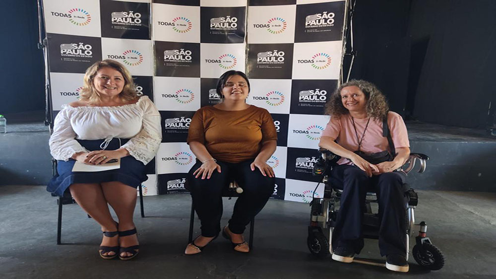 Imagem colorida do Encontro de Mulheres em Capivari – Todas in-Rede. A Secretária Arácelia Costa e outras mulheres, sentadas, em frente ao painel do programa no evento.