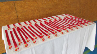 Imagem colorida. Uma mesa com toalha branca. Sobre a mesa medalhas com o logo do SRLM e texto em Braille.