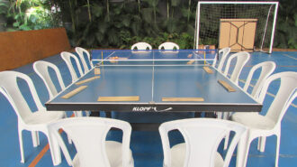 Imagem colorida da quadra do SRLMH. Uma mesa de tênis e ao redor cadeiras. Sobre a mesa raquetes.