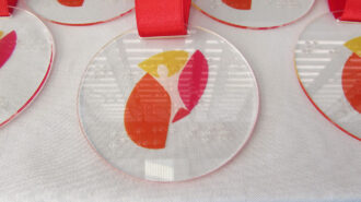 Imagem colorida da medalha. O logo do SRLM e o texto em braille em um circulo de acrílico.