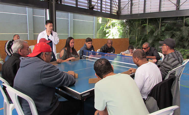Fotografia colorida da quadra do Serviço de Reabilitação Lucy Montoro Humaitá. Pessoas sentadas ao redor de uma mesa de tênis, jogando Takkyu Volley. Ao lado, de jaleco, o árbitro.